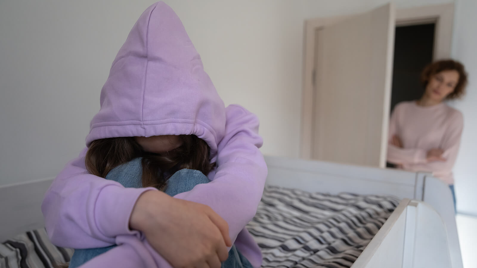Image of a young girl sitting on her bed with a hood covering her face with her mom looking in the doorway. Learn how anxiety in teens can affective their mental health and find support with teen counseling in Macon, GA.