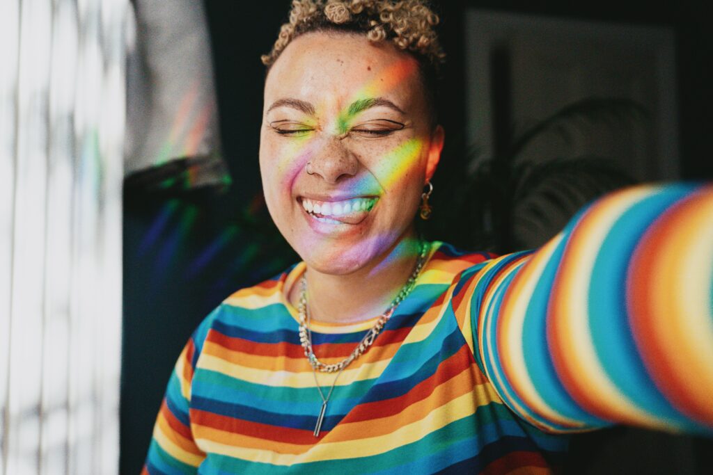 Photo of an African American woman wearing a striped rainbow shirt and smiling with her eyes closed while a rainbow reflection shines on her face. Begin to work through your past trauma with the help of trauma intensives in Macon, GA.
