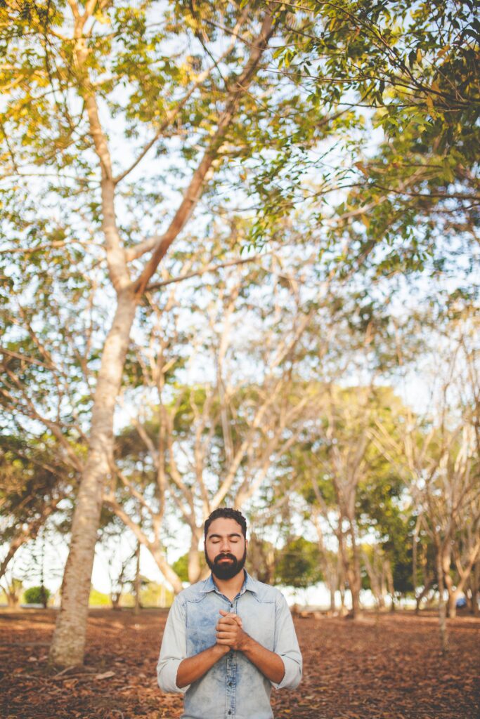 Image of a bearded man standing under trees with his eyes closed and hands clasped together. Work through your trauma symptoms with the help of a trauma intensive in Macon, GA.